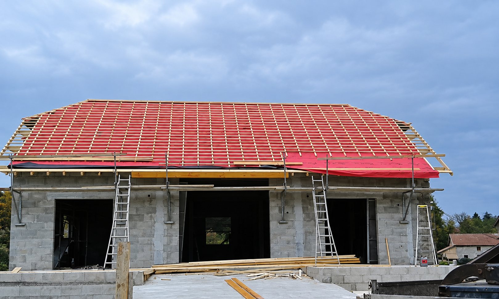 Maison en parpaings en construction dont la charpente en bois est recouverte d'un écran rouge, avec des échelles et des échafaudages devant la façade.