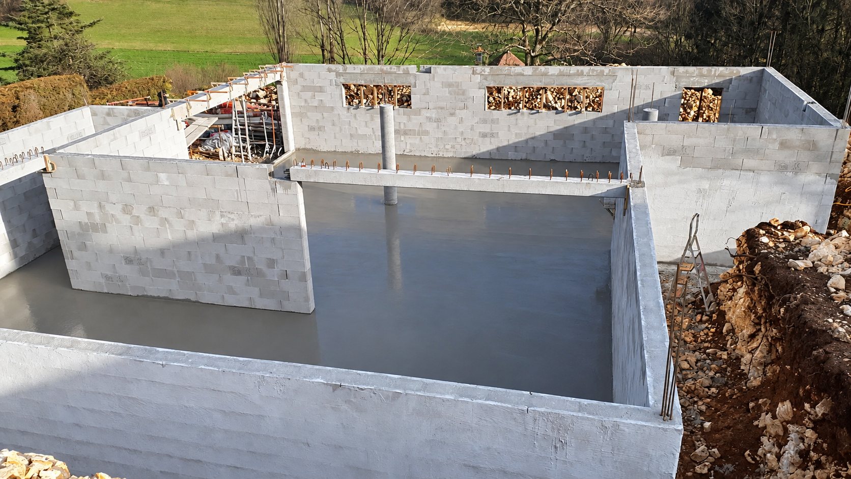 Vue en hauteur d'un chantier de maison dont les murs en parpaings entourent une dalle de béton fraîchement coulée, au milieu d'un paysage de campagne.