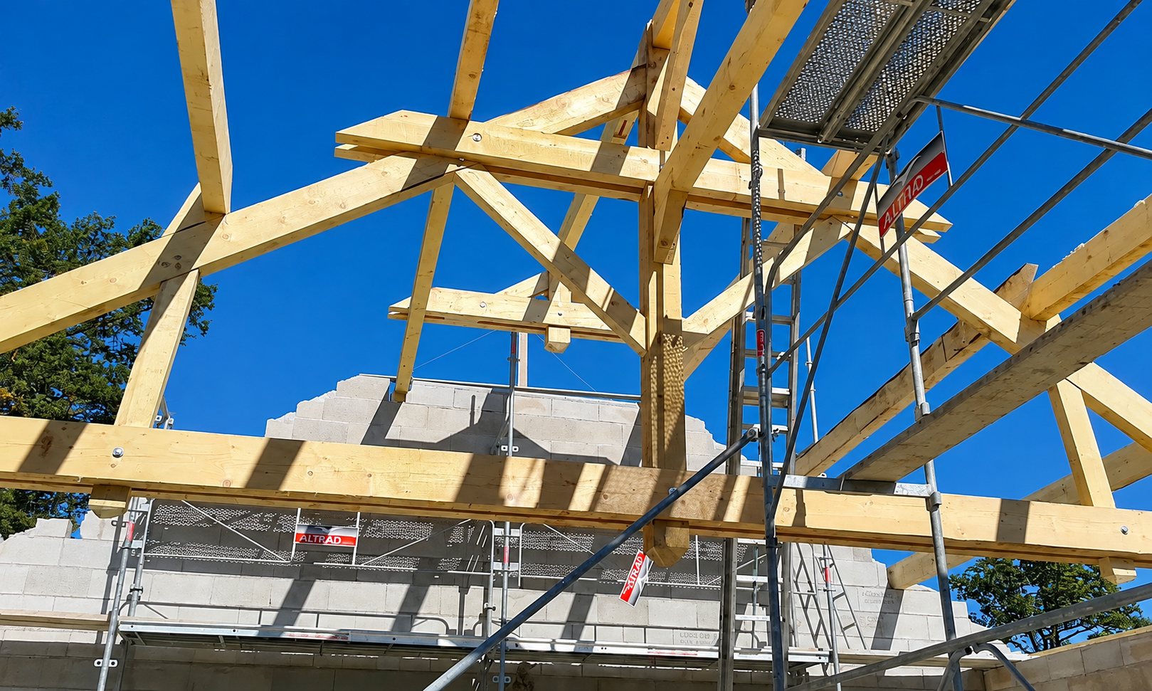 Charpente en bois neuve en cours de montage au-dessus de murs en parpaings, entourée d'un échafaudage sous un ciel bleu.