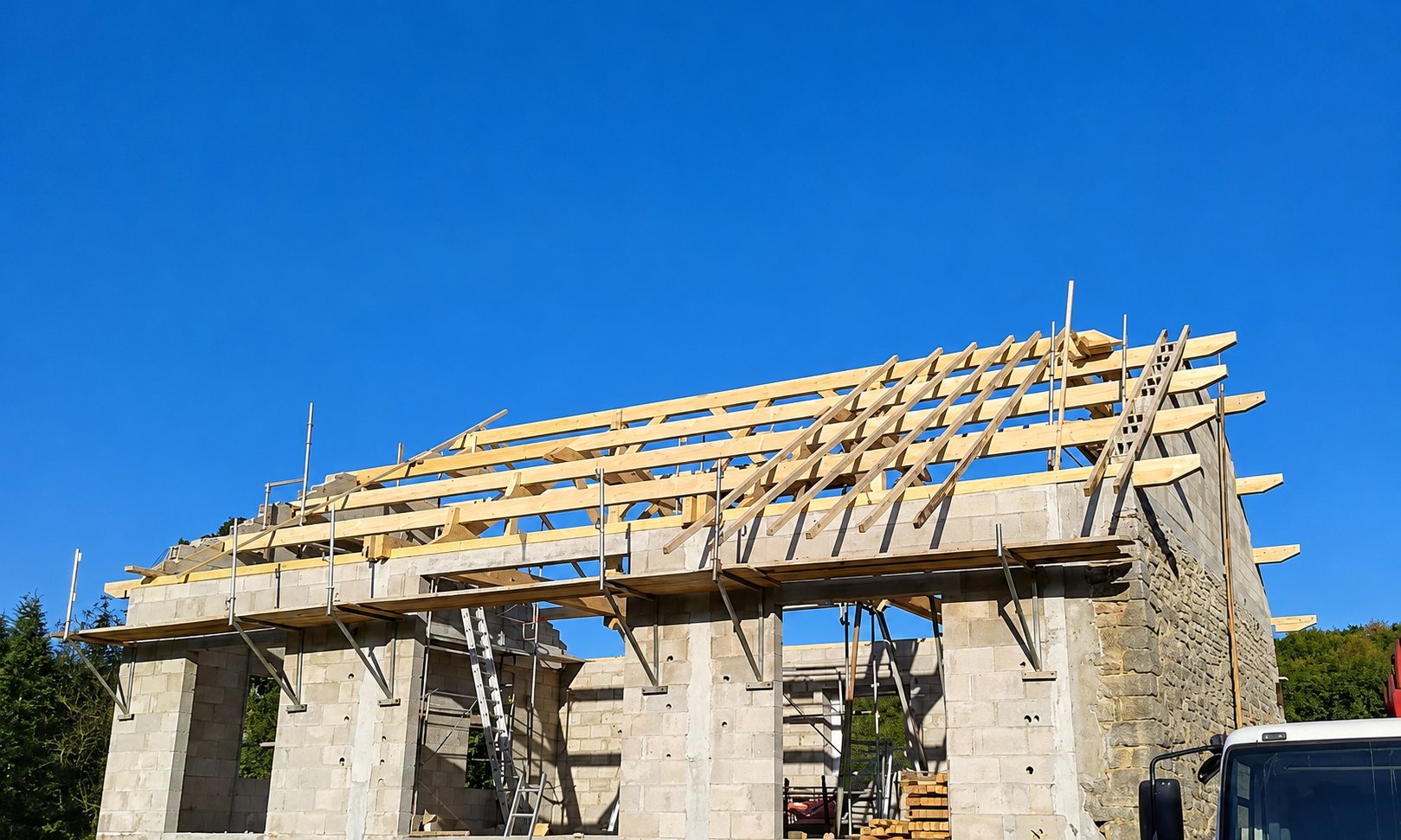 Une charpente en bois en cours de pose sur les murs en parpaings d'un bâtiment en construction, sous un ciel bleu.