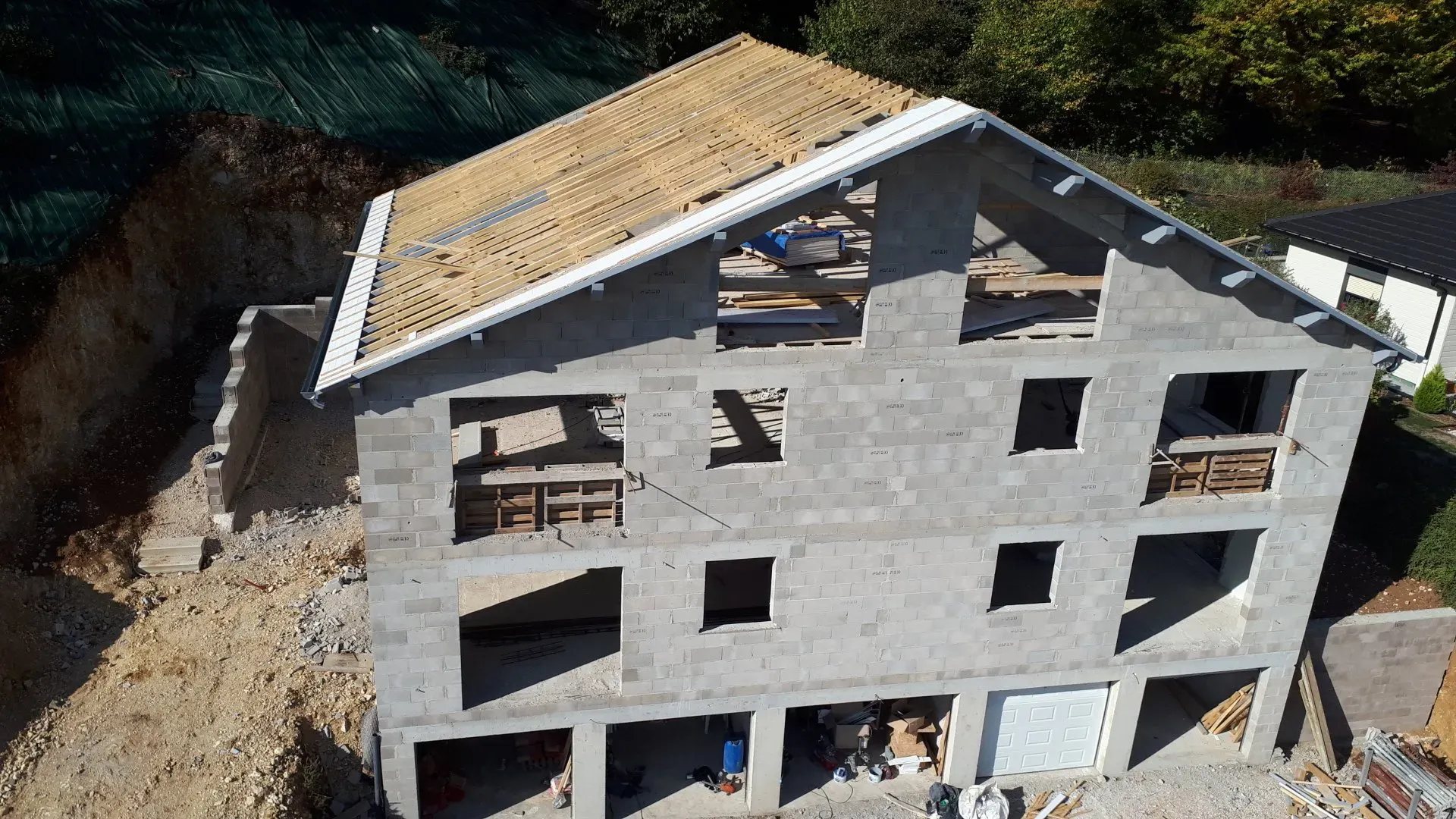 Vue aérienne d'un chantier de maison en blocs de béton dont la charpente en bois est posée, avec des garages au rez-de-chaussée et un terrain terrassé.